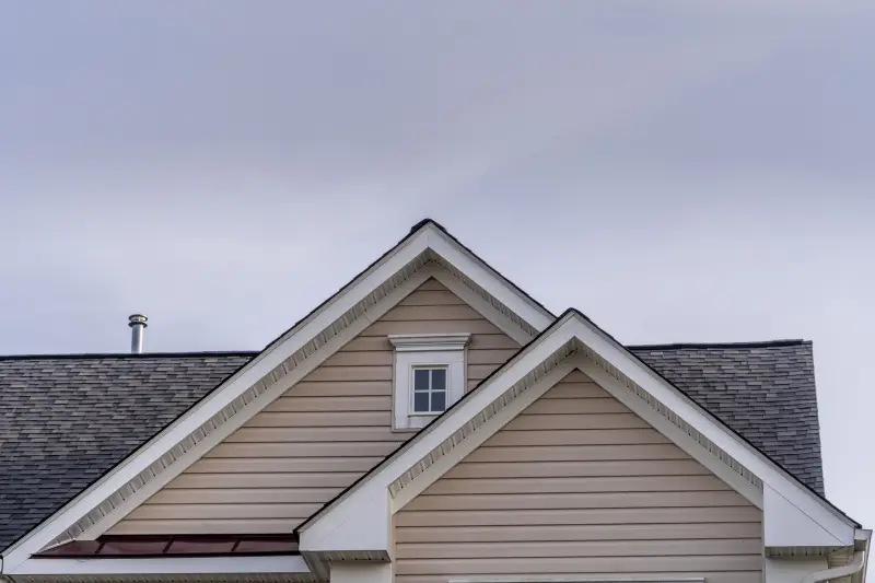 Beige horizontal vinyl siding, white decorative frame attic window reverse double gable close up on luxury single family residential home. Square single pane gable window. Roof line of traditional home with one gable running one way reverse gables running the other.